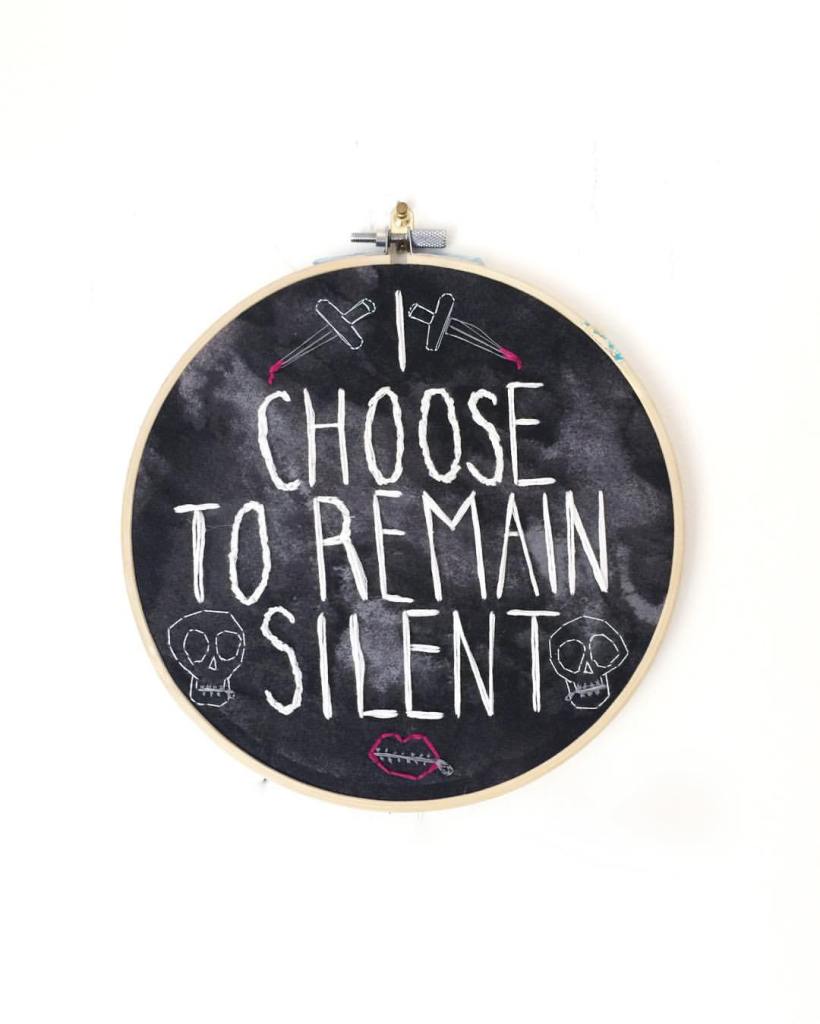 An embroidery hoop with needlepoint showing the words I Choose to Remain Silent, bordered by daggers, skulls, and a mouth zipped shut.